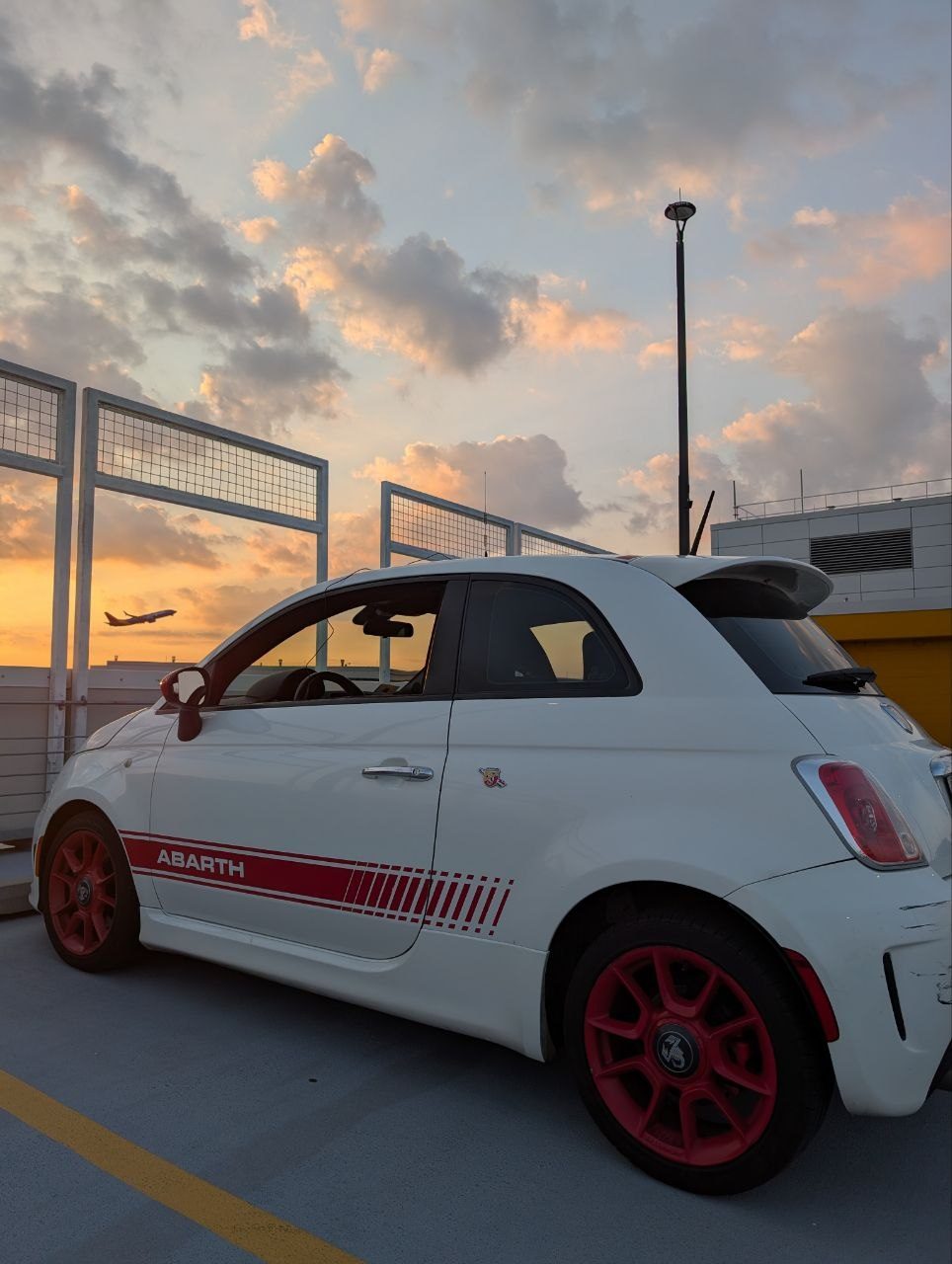 A white Fiat 500 Abarth with a magnetic antenna on its roof, parked on the top floor of the Midway garage at sunset, a jet climbing out behind it