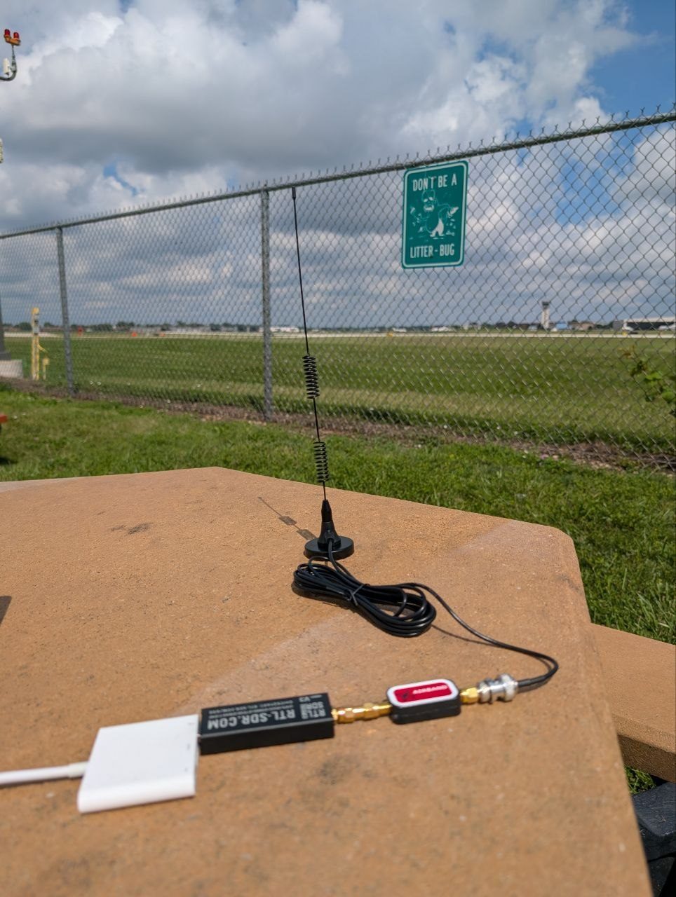 A magnetic-mount scanner antenna standing on a concrete barrier at the Chicago Executive viewing area, with the airfield fence and runway behind it
